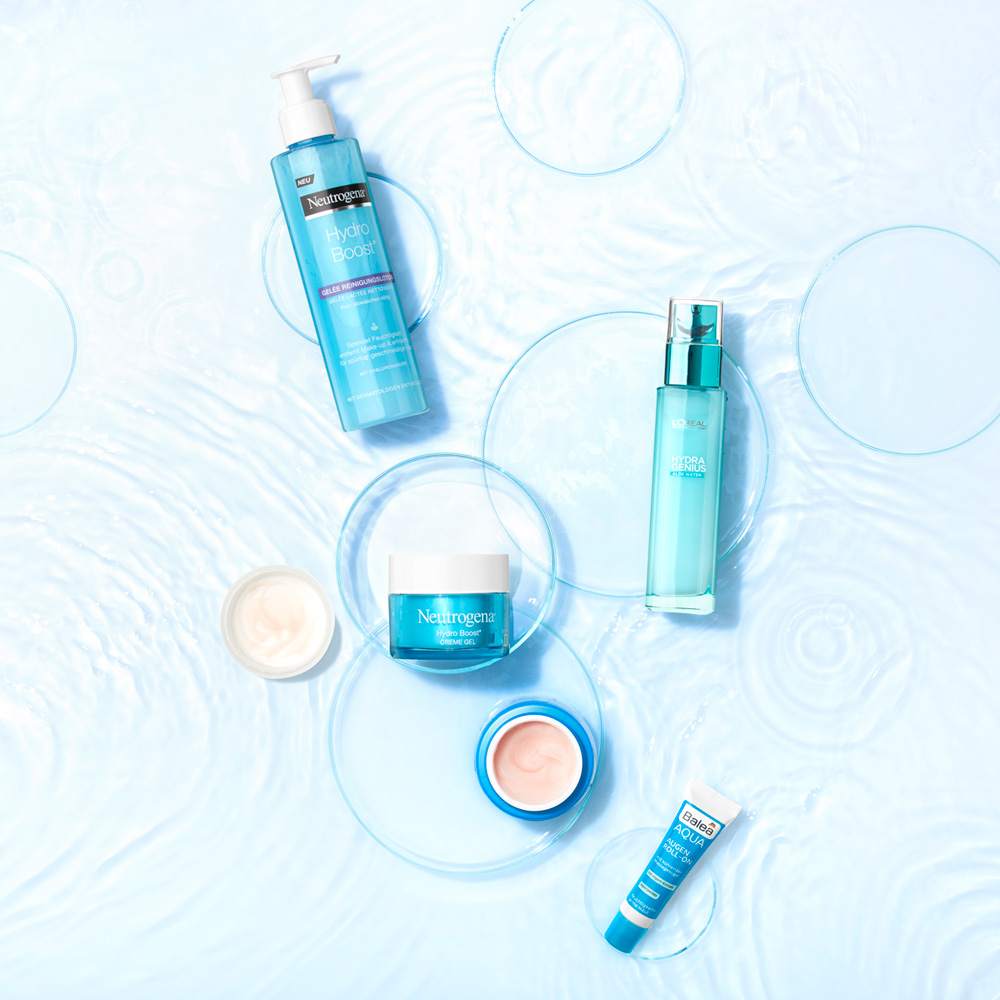 Aufsicht auf 5 unterschiedliche Kosmetikproduktbehälter auf einem Untergrund aus ringeschlagendenm Wasser und Petrischalen