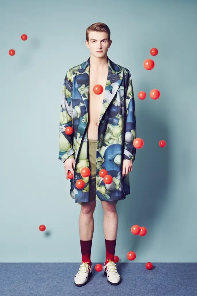 A male Model wearing a acket and Bermuda Shorts, socks and white Slippers. Red plastic balls are flying around infront of him.